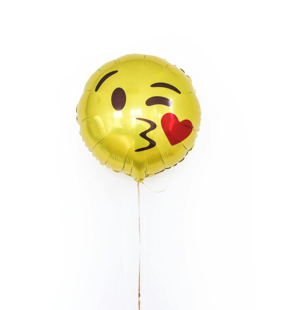 Golden glossy round shaped balloon with smiley kissing heart face filled with helium arranged on white background isolated in studio