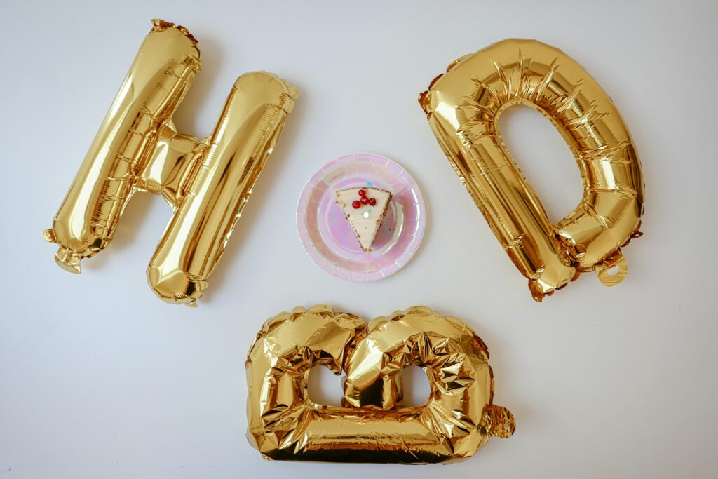 A festive birthday setup with a cake slice and shiny gold foil balloons spelling 'HBD'.